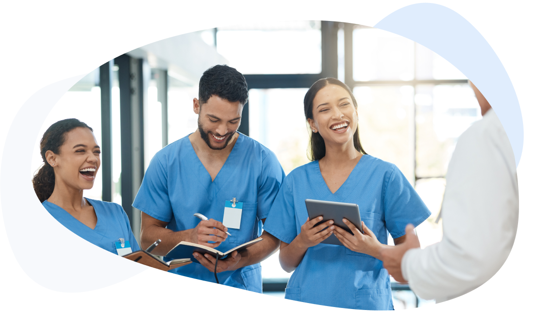 Three smiling medical staff in blue scrubs take notes in a notebook and on a tablet.