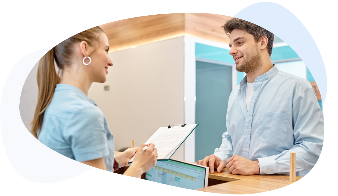 Receptionist hands a form to a man at the desk; a visit schedule is visible on the monitor.